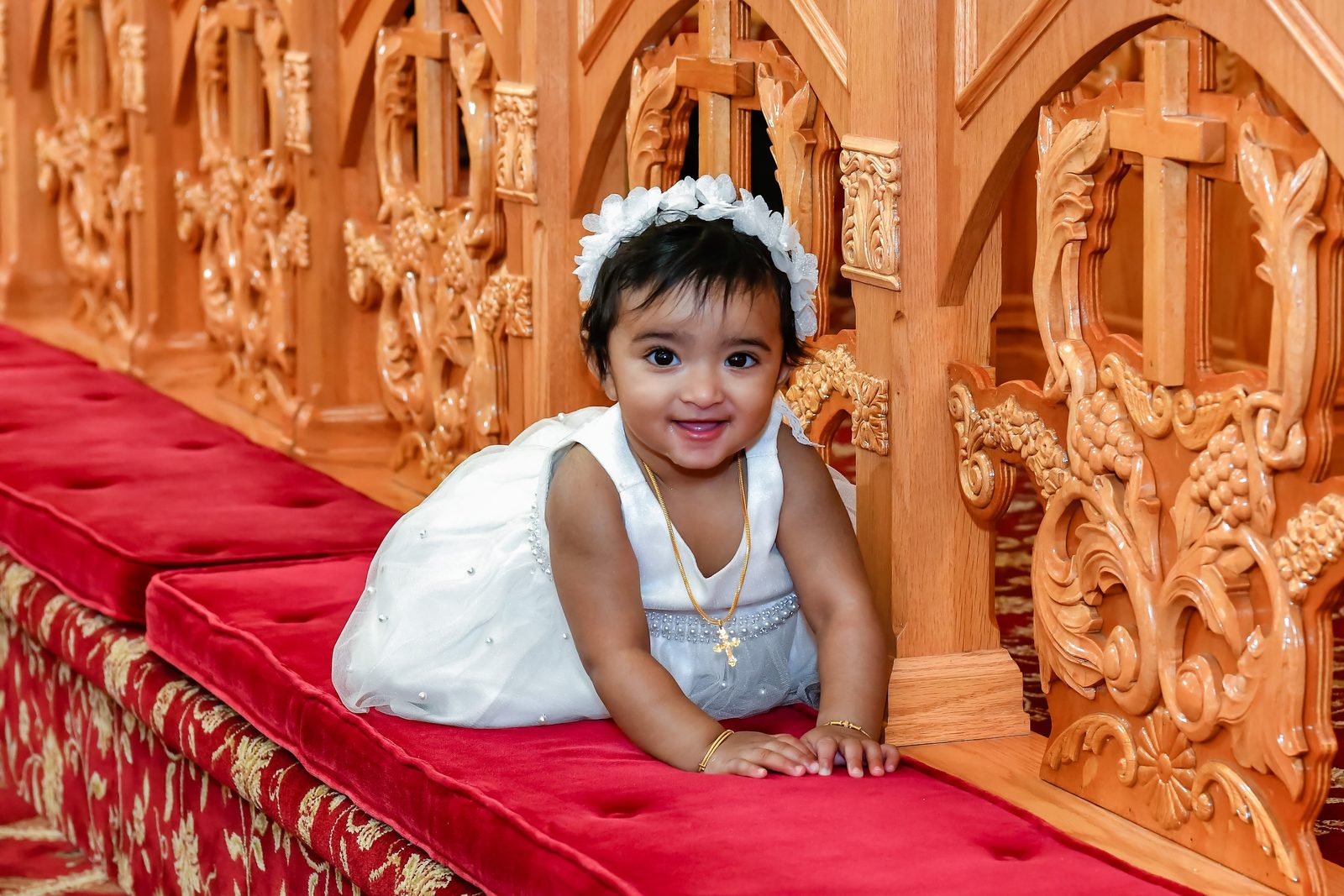Baby in a white dress smiling on a red church pew