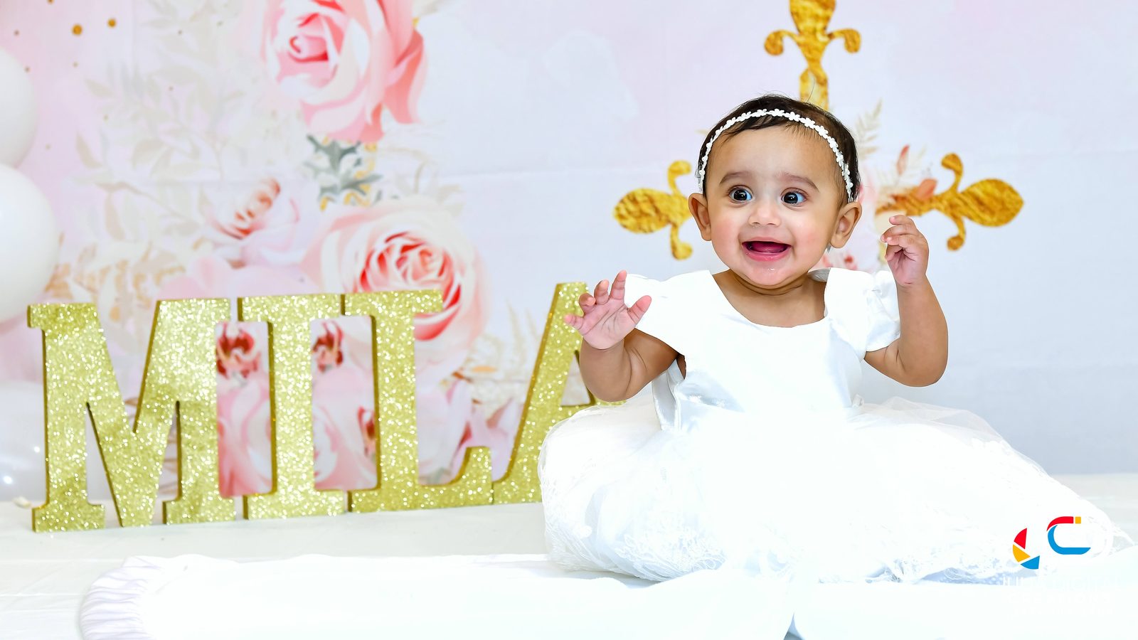 Smiling baby in a white dress and tiara beside gold name letters
